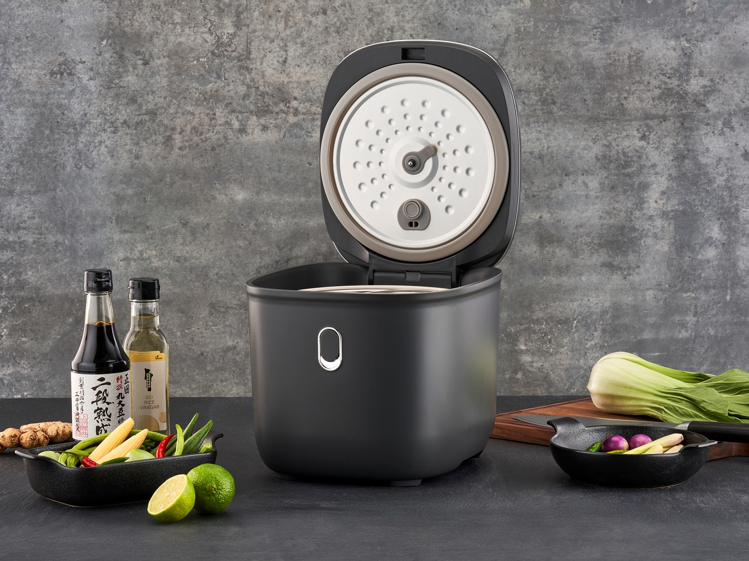 Offener Reiskocher in modernem Design, umgeben von frischen Zutaten und asiatischen Gewürzen.